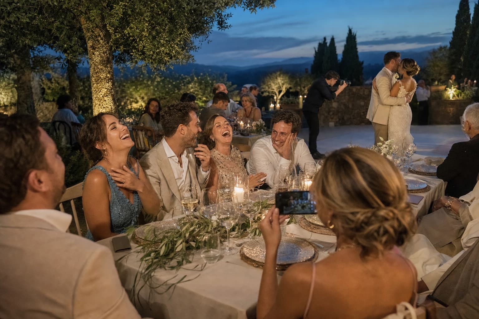 Invitada fotografiando discretamente las risas espontáneas de una mesa mientras el fotógrafo profesional trabaja al fondo